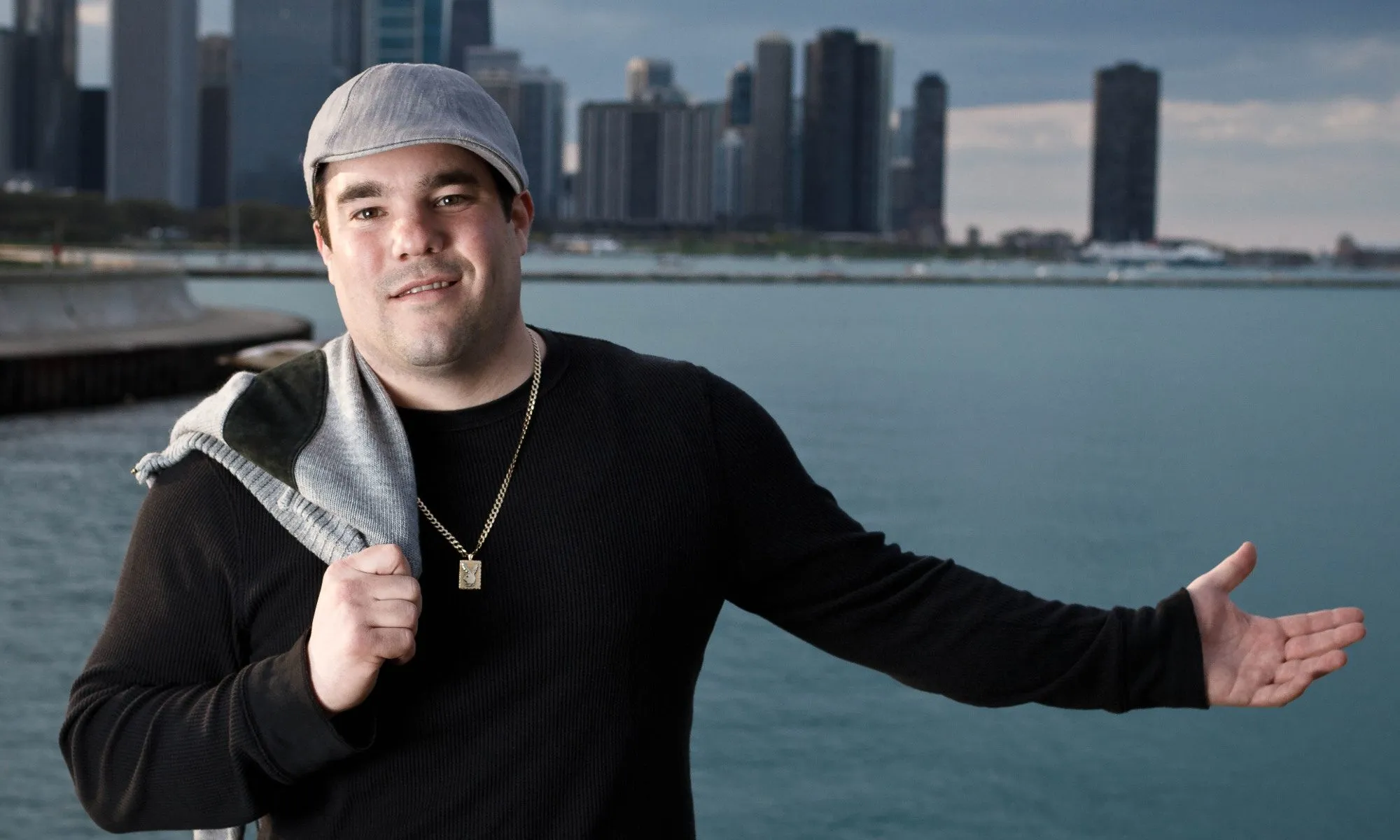 Vinnie dressed in a black shirt, one arm outstretched, the other draping a gray and black sweater over his shoulder. Lake Michigan and the Chicago skyline are visible in the background.